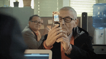 Video gif. An old man holds a flip phone, spinning it around in his hands. He squints at it with confusion. A man behind him watches him struggle and then looks away, itching his ear as if experiencing second hand embarrassment