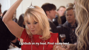 Video gif. A woman wearing a dress in a crowd, holds a champagne glass above her head as she states manically, "Light Bulb on my head. Pinot on my head."