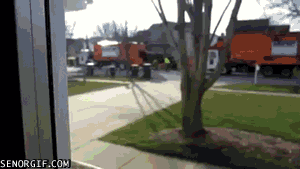 Video gif. We zoom in across a street where two garbage collectors in neon yellow vests are playing a spirited game of foosball beside their orange truck.
