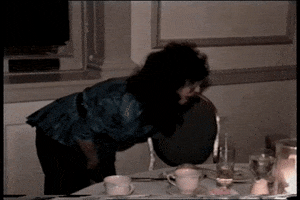 Video gif. Home video of a woman struggling to sit in her seat at a fancy dinner. She falls out of her chair, her feet flying into the air as she disappears from view. A woman sitting at the table watches her fall down and bursts into hysterical laughter.
