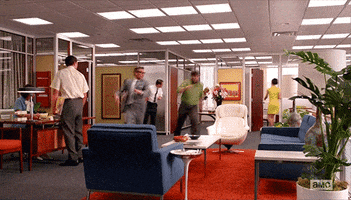 TV gif. A scene from Mad Men where two workers are running through the office, hopping over sofas and tables in a mad dash.