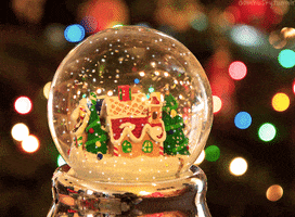 Photo gif. Snow falls in a snow globe over a gingerbread house surrounded by pine trees, as it sits in front of a Christmas tree in the background. 