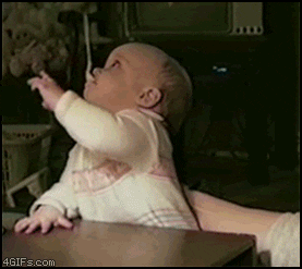 Video gif. A baby can't figure out how to take the spoon out of their mouth, reaching for and missing it with both hands.