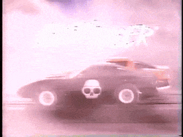 Ad gif. Retro 80s TV commercial of the Ghost Racer shows a black miniature toy car with a white skull racing an orange car through a smokey scene.