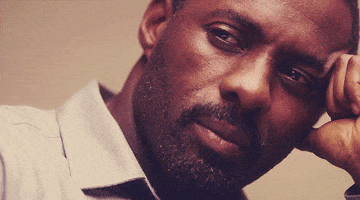 Celebrity gif. Idris Elba is leaning his head on his hand and he slowly smiles.