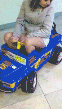 Video gif. Woman sits in a toy car that she’s too big to fit in properly.