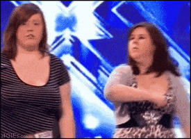 TV gif. Two women are performing on the X Factor. One of the women deliberately hits the other woman in the face and she looks at the judges, shocked.