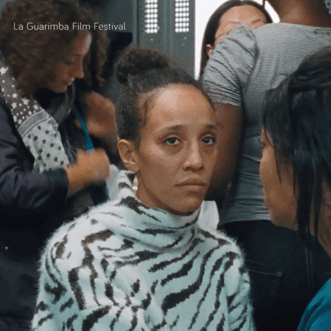 Video gif. Woman sits amongst a group of people who are talking, laughing, and moving around. Unlike the people around her, she sits with a blank, tired expression on her face. She looks into space with heavy bags under her eyes.