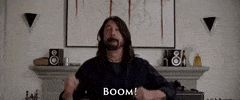 Celebrity gif. Dave Grohl of the Foo Fighters stands in front of a living room mantelpiece with his guitar strapped to him. He leans back and throws his arms up as he exclaims: Text, "Boom!"