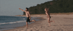 Movie gif. On a beach, Olivia Wilde as Kate in Drinking Buddies gallops after Jake Johnson as Kyle, who trips and falls once he reaches the water.
