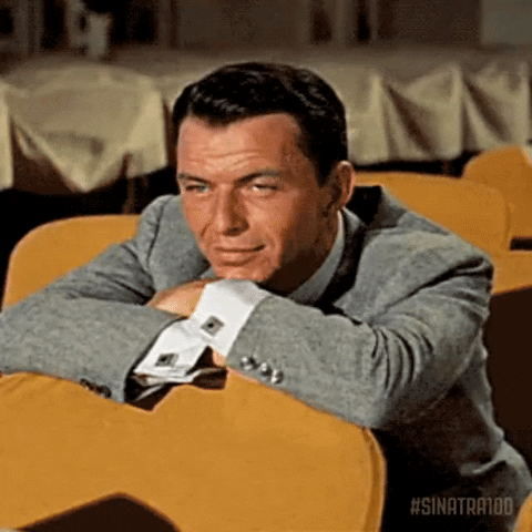 Celebrity gif. A thoughtful Frank Sinatra leans against the back of a chair and smiles, blinking.