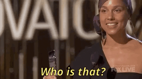 Celebrity gif. Alicia Keys holds an award on a stage. Her hair up in a wrap as she turns with a smile. Text, "Who is that?"