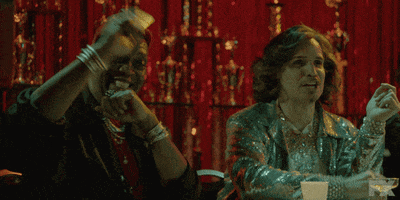TV gif. Two men on Pose sitting behind a table, in front of a set of large trophies, snapping their fingers and smiling with approval.