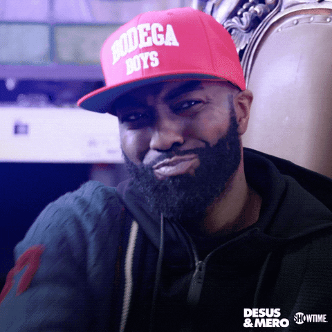 TV gif. Sitting in a fancy chair, wearing a red "Bodega Boys" cap and a sweater over a black hoodie, Desus Nice of Desus and Mero smirks and points to us.