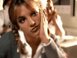 Music video gif. From the video for "Baby One More Time," Britney Spears is dressed in a school uniform, sitting at a desk, resting her head in her hand and looking bored until she cracks a smile.