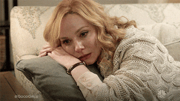 TV gif. Christina Hendricks as Beth on Good Girls cradles a throw pillow as she lies on the arm of a couch, staring off blankly and crying.