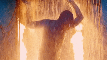 Music video gif. From video for Ricky Martin's Perdóname, a man stands backlit underneath a cascading spray of water, appearing to seek refuge and holding his arm up while a large fire blazes behind him.