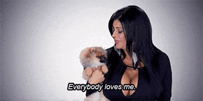 Reality TV gif. Ang Raiola on Mob Wives holds her little pomeranian puppy in her arms. She looks at us and shrugs with a cheeky smile as she says, “Everybody loves me.” Three hearts appear above her puppy’s head