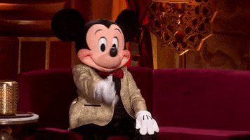 Disney gif. Person wearing a genuine Mickey costume sits on a red velvet couch wearing a gold jacket and red bowtie. He shakes his head and puts a white gloved hand to his face like he's amazed or embarrassed.