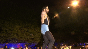 Video gif. Man wearing black bodysuit and tights, denim shorts, and rainbow suspenders dances flamboyantly while police lights flash below.