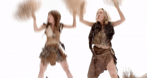 Gif of two women dressed like cave women waving pom poms made from tumbleweed