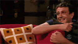 TV gif. Jason Segel as Marshall on How I Met Your Mother kneels behind a chair, embracing a pillow. He looks up with a goofy smile, completely enamored with what he’s looking at. He hides half of his face behind the pillow and looks up with loving eyes.