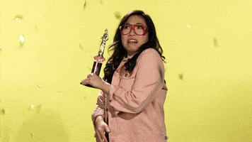 Celebrity gif. Awkwafina holds a trophy and shudders as she sees confetti flying around her. Then, she embraces it and smiles and waves at us while playing with the floating confetti.