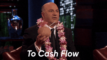 Reality TV gif. Kevin O'Leary on Shark Tank wearing a lei and holding a drink in a coconut. He toasts us and says, "To cash flow!"