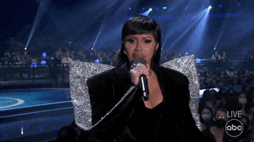 Celebrity gif. Cardi B is performing at an awards show and she holds a microphone and pouts widely while throwing up a hand to stop the crowd's noise.