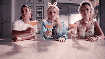 TV gif. Billie Lourd as Chanel #3, Lea Michele as Hester, and Abigail Breslin as Chanel #5 from Scream Queens clink martini glasses in a somber cheers.