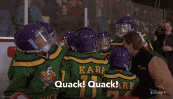 Movie gif. The Mighty Ducks team and Emilio Estevez as Gordon Bombay stand in a circle and hold their hands in the middle and chant, “Quack! Quack!”