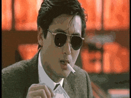 Video gif. A man in a suit wearing sunglasses lights a cigarette in his mouth with a $100 bill that is on fire.
