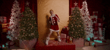 Holiday gif. Ed Sheeran in a Santa romper and white boots stands on a stage that's actually a humongous red gift box and slowly sways side to side while grabbing the ends of a thin, fuzzy white scarf. A wrapped present topples over from his foot. He's surrounded by a symmetrical display of green and white Christmas trees decorated with ornaments while high-rising stacks of presents are cluttered in the background.
