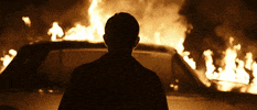 Movie gif. With his back to us, Jack Roth as Danny in Us and Them gazes upon the scene of a car on fire, with flames and smoke lapping in slow motion.