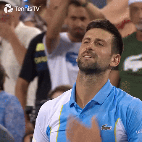 Sports gif. Novak Djokovic walks across the tennis court, ripping his shirt off as he screams in triumph with fans clapping in the stadium behind him. TennisTV logo in the corner.