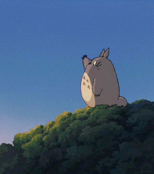 Anime gif. Totoro from My Neighbor Totoro stands on a treetop and waves offscreen.