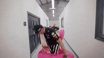 Sports gif. Racing driver Max Verstappen stands in a hallway with a towel flung over his shoulder, pulling a pink flamingo inflatable up around his waist, then waddles away in flippers.