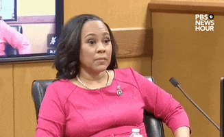 Political gif. Fulton County District Attorney Fani Willis sits in a courtroom wearing a hot pink top with a microphone in front of her. She leans back in her chair in a relaxed, self-satisfied manner, winking and smirking at someone offscreen.