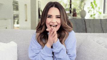 Video gif. Woman is ecstatic and squeals while she puts her hands to her cheeks and sits back in the couch, curling up in anticipation.