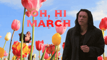 Meme gif. Tommy Wiseau as Johnny in The Room stands in front of a background of tall, colorful tulips. He tips his head up with a blank expression on his face as he says, “Oh, Hi March.”