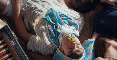Music video gif. From the Once in a While video, a member of Timeflies lies on the floor, wearing a baby bib and bonnet, as he sucks on a yellow pacifier.