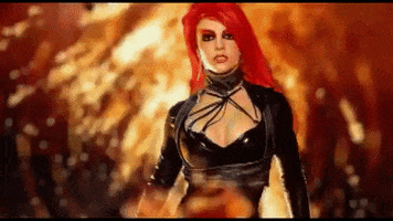 Music video gif. Britney Spears wears a black leather outfit, walking from a fire as her long red hair flies around her, in the video for her song "Toxic."