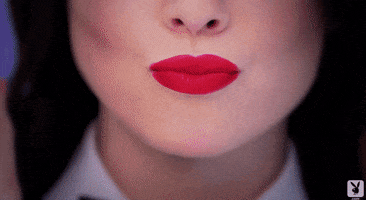 Video gif. We see a shot of a woman's nose and cherry red lips. She sends us a kiss before breaking out into a smile.