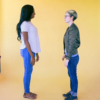 Video gif. Two women mirroring each other bounce upwards smiling and lift one leg playfully, giving each other two high fives.