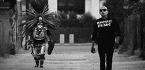 Video gif. Black and white video shows two people walking towards us in slow motion, one dressed in an ornate feathered serpent costume and the other dressed in black, wearing a shirt that reads "Brown pride."
