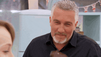Reality TV gif. Paul Hollywood on the Great British Bake off looks at someone with a cheeky smile as he rubs his chin almost like he’s jokingly suspicious of something. 