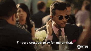 TV gif. Utkarsh Ambudkar as Malcolm on White Famous talks into an earpiece as he sits outside at a restaurant wearing aviator glasses, smiling and crossing his fingers, as he says, "Fingers crossed; pray for me on that."