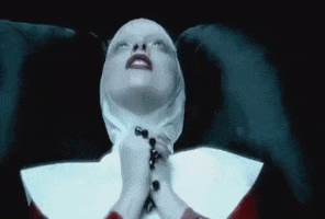 Music video gif. Lady Gaga in Alejandro is dressed as a nun. She holds a rosary in her hands as she lays on a black leather couch. She thrusts her hands in the air and says, "Alejandro," before bringing the rosary back to her lips.