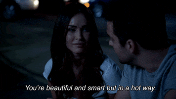 TV gif. Jake Johnson as Nick and Megan Fox as Reagan in New Girl. They're sitting on a curb and Nick tells Reagan, "You're beautiful and smart but in a hot way," and it makes her smile.