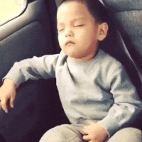 Video gif. In a car, a sleeping toddler wakes to the sound of music and begins to dance, bobbing his head in excitement.